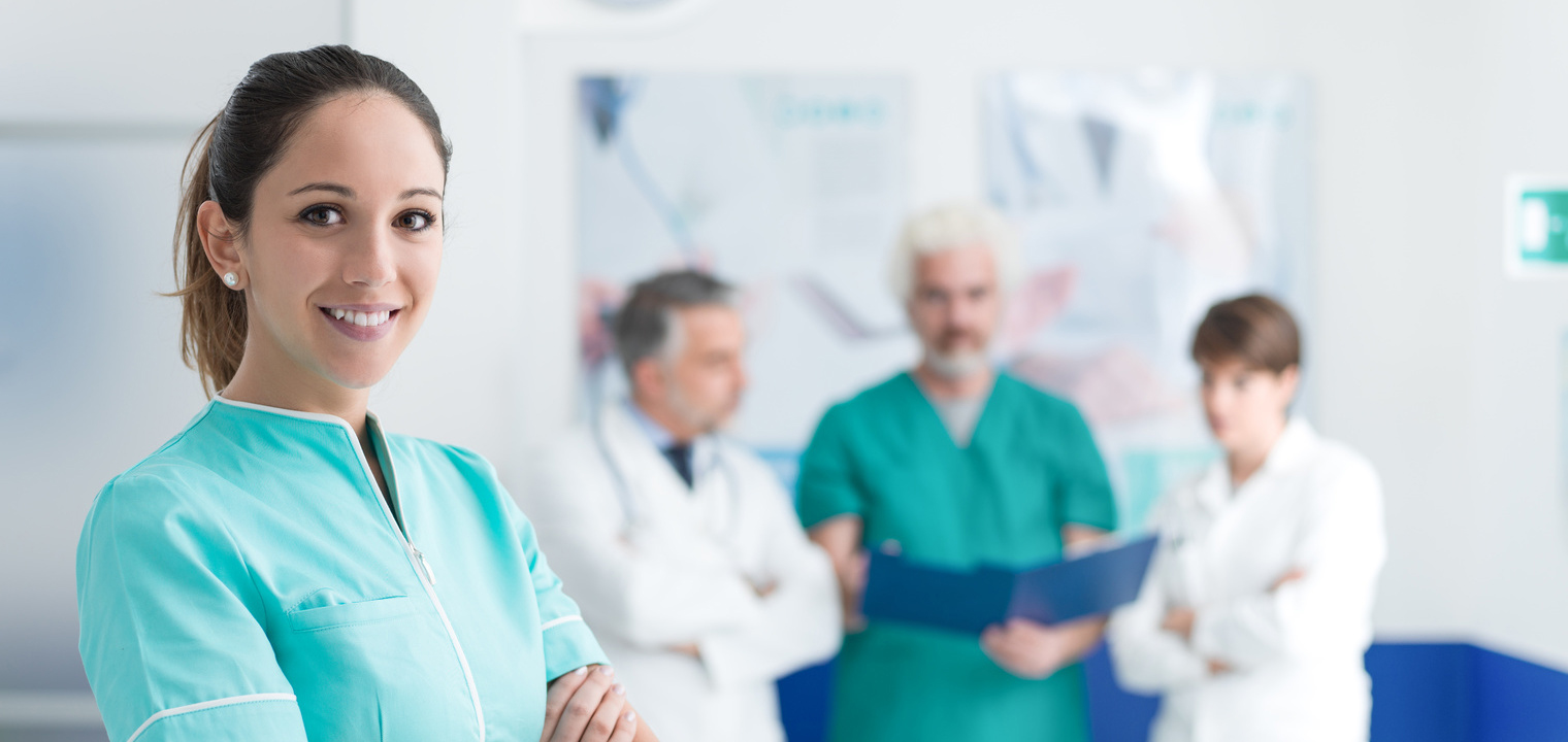 Young nurse posing at the clinic with arms crossed and medical team working on the background